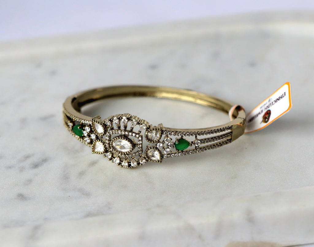A vintage-style gold bangle openable bracelet with a detailed filigree centerpiece featuring clear stones and green emerald-cut accents.