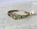 A vintage-style gold bangle openable bracelet with a detailed filigree centerpiece featuring clear stones and green emerald-cut accents.
