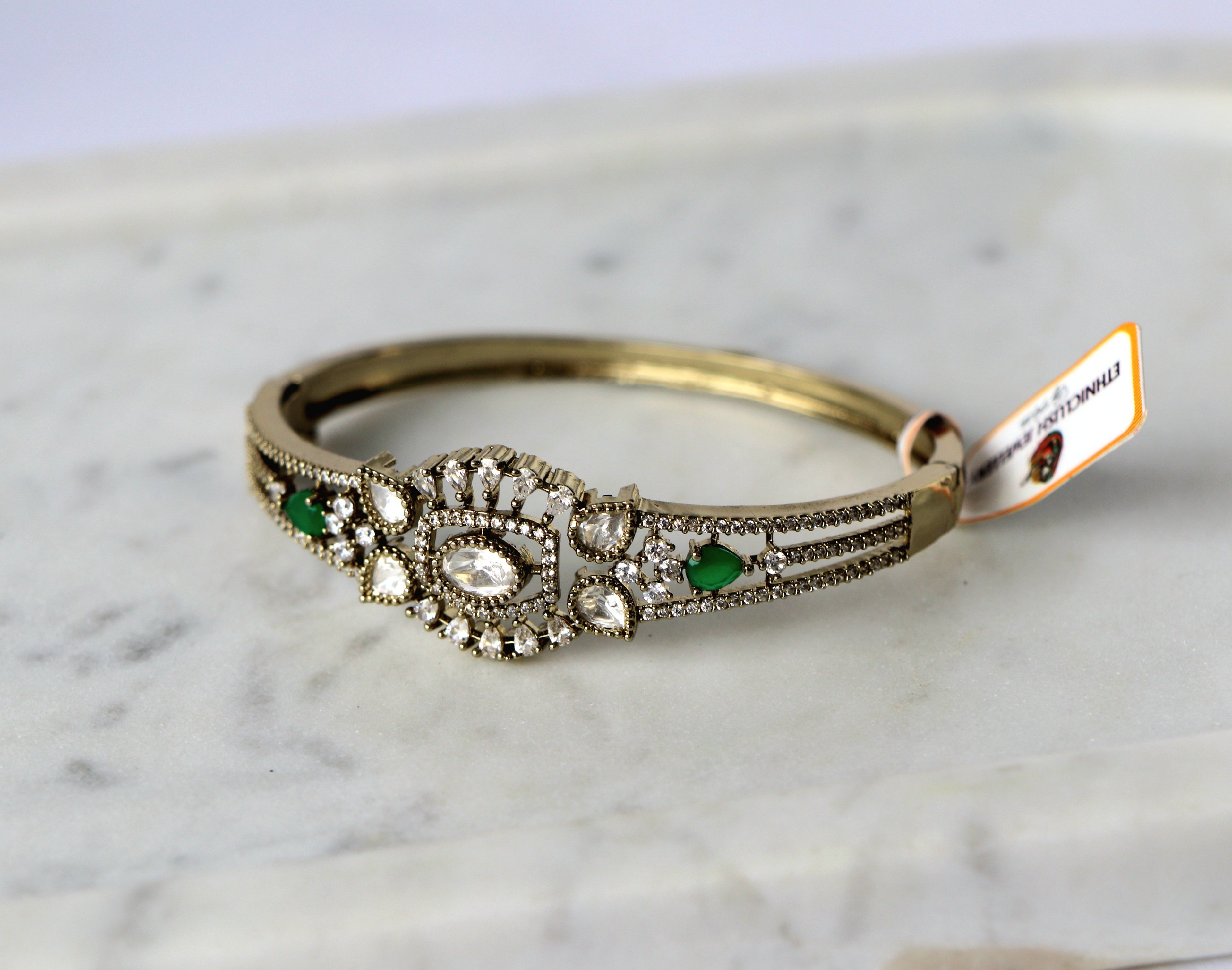 A vintage-style gold bangle openable bracelet with a detailed filigree centerpiece featuring clear stones and green emerald-cut accents.